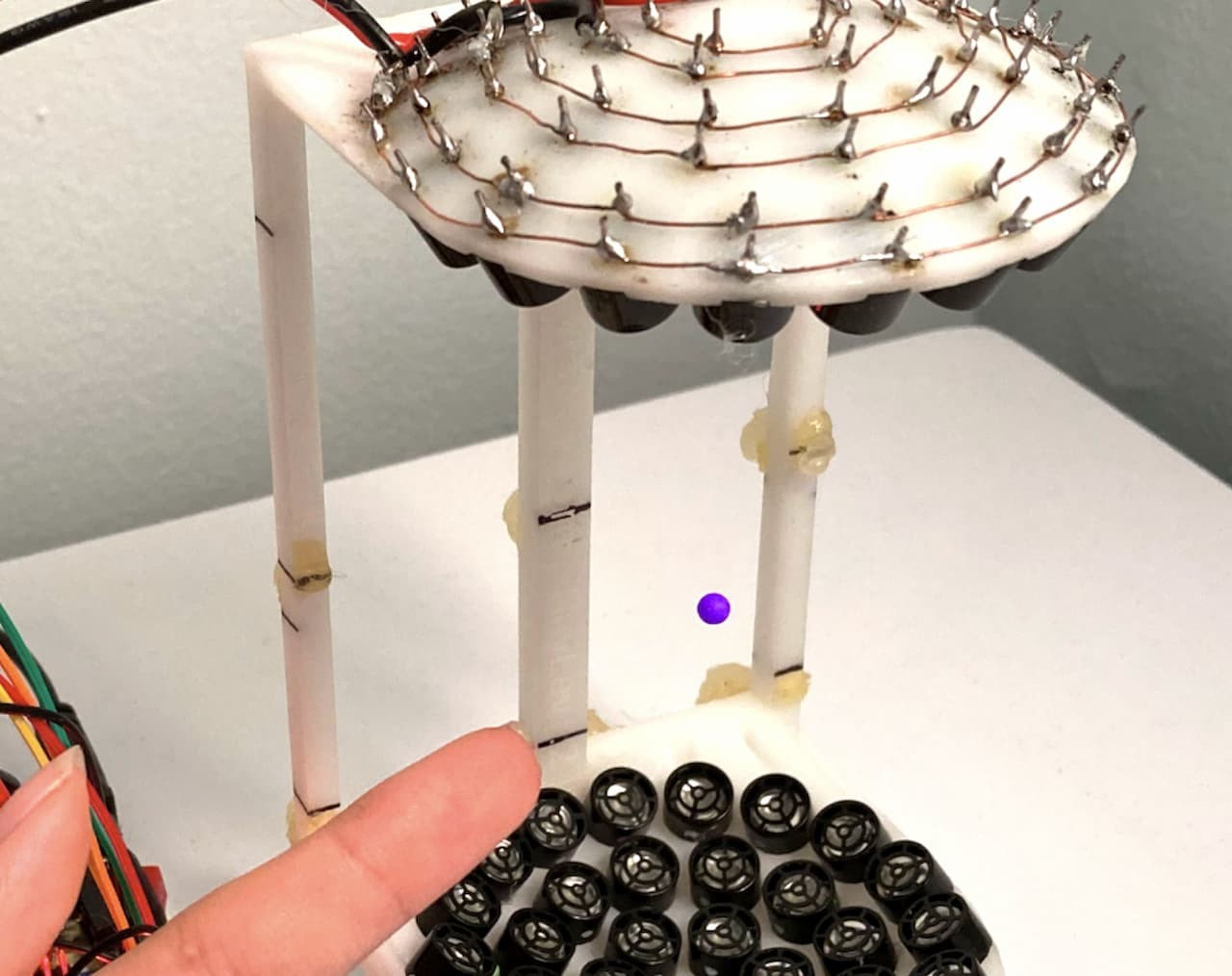 Ένα σφαιρίδιο (μωβ) αιωρείται στον αέρα μέσω ηχητικών κυμάτων που εκπέμπονται από (μαύρα) κυκλικά ηχεία, τοποθετημένα σε ένα τρισδιάστατα εκτυπωμένο πλαίσιο ύψους έξι ιντσών. Πηγή: Κέντρο Έρευνας Μαλακής Ύλης του NYU (NYU's Center for Soft Matter Research)