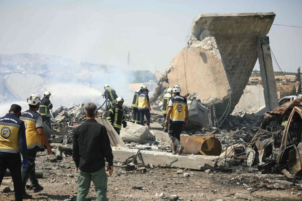 Διασώστες των Syrian White Helmets επιθεωρούν τις ζημιές, ενώ πυροσβέστες επιχειρούν για την κατάσβεση της φωτιάς στο σημείο της έκρηξης σε προσωρινή αποθήκη όπλων στην πόλη Σαρμάντα, στην επαρχία Ιντλίμπ, την Τετάρτη. Φωτογραφία: AFP